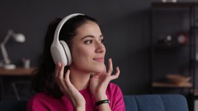 Beautiful girl enjoying music on couch at home. Close up portrait female listening to music in modern headphones. Pretty young woman dancing and listening music with white headphones.
