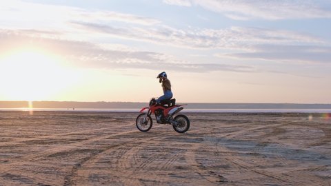 Girl biker, quickly rides along the sandy coast of the lake, at sunset. A stuntman doing stunts on a motorbike in motion.