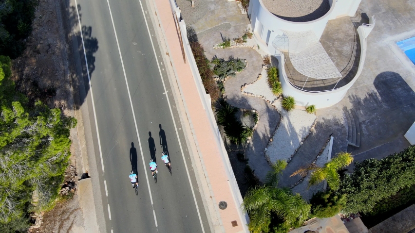 Artistic birdseye drone footage of three team cyclists riding along a straight road in Calpe, Spain, on a warm morning with long shadows, and traditional terracotta roofed buildings lining the road