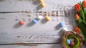 4k video. Multicolor Easter eggs rolling on white wooden table with tulips. Traditional Spring composition Easter elements flowers and eggs for springtime. Spring decorations concept