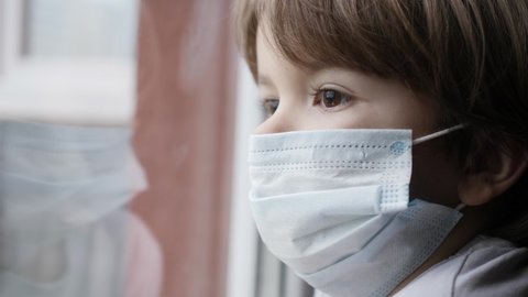 Little Child Boy With Protective Mask, Sad Looking Through Window Worried About Covid-19 Lockdown. Kid In Protection Mask Looking Out Window Home. Quarantine Coronavirus Social Distancing