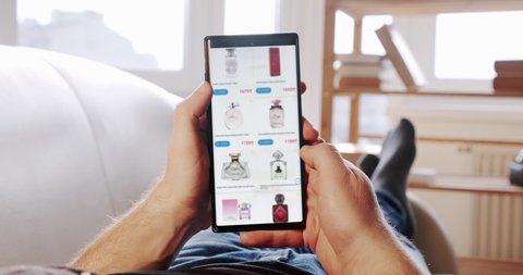 Man at Home Lying on Couch Orders perfumes In blured Online Store using delivery app on a Smartphone. POV