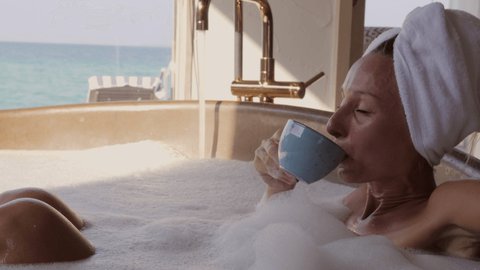 Young woman enjoys a cup of tea wile taking a relaxing bath. Female taking a bath in luxury villa overwater with stunning sea view 
