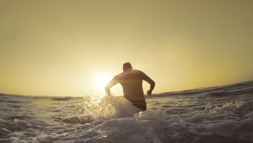 Man running on the beach in shallow water and jump swimming at sunset, slow motion