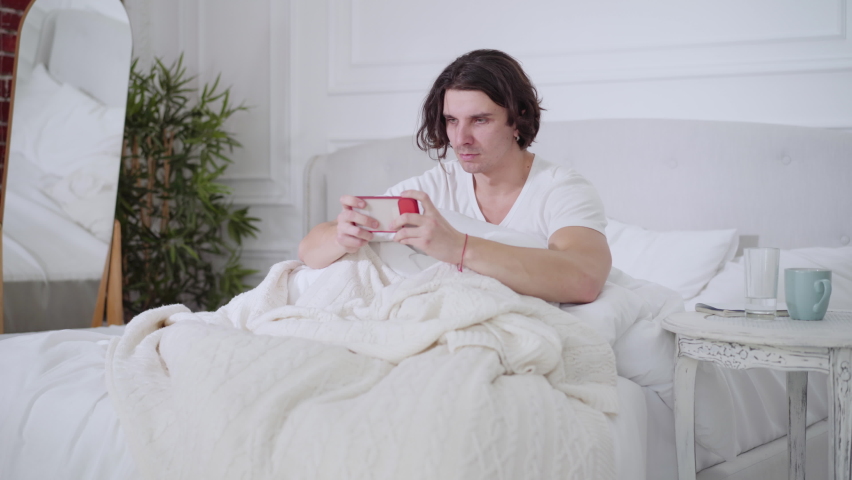Concentrated young brunette man gaming online on smartphone and making victory gesture sitting in bed in the morning. Portrait of device addicted Caucasian gamer in bedroom at home.