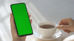 Woman Hand Cup of Hot tea and Using Smartphone Watching Green Screen Top View. Smartphone with Green Mock-up Screen Business Concept.