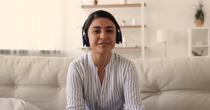 Happy beautiful millennial indian mixed race woman in earphones looking at camera, talking discussing working issues distantly, giving educational lecture online, sharing knowledge via video call app.