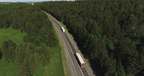 Two Semi Truck with white trailer and cab driving, traveling alone on dense flat forest asphalt straight road, highway top view follow vehicle aerial footage. Freeway trucks traffic