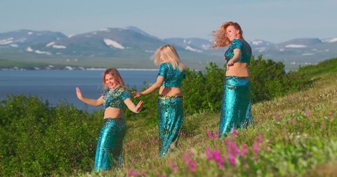 Women dance in exotic costumes and do torso turns with flowing hair. Shot in the general shot against the background of green grass, water and mountains, on a sunny day. Slow motion