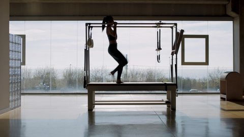 Tracking shot in slow motion of Fit female instructor hanging from the pilates machine, woman working out on the Pilates Trapeze Table in a big Fitness Studio with wide windows and street views.