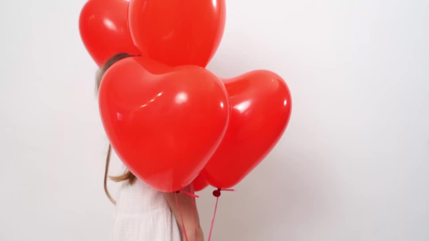 Funny beautiful little girl with red balloons smiling and folds palms in front of him near the white wall. celebration and fun. Surprise for the child. Valentine
