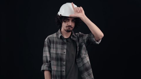 Young engineer holding his fingers near his forehead, showing the sign of loss with sarcasm. Young man with curly hair wears a special helmet on his head, dressed in a plaid shirt, on a black