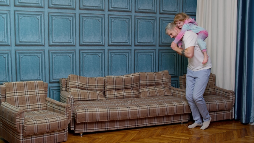 Happy carefree family dad and child kid daughter dancing, playing piggyback, running in living room at home. Funny teen girl riding on father, having fun, fooling around during morning on holidays