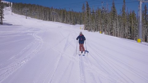 Skier on a groomed ski resort trail using her ski poles to speed up while passing a ski lift