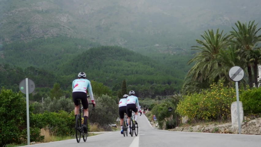 Realtime low angle footage of three team cyclists riding down a straight road towards a forested hilly region in Calpe, Spain