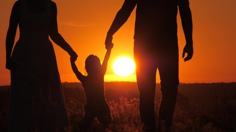 The child plays with dad and mom on field in light of sunset. Happy family, little daughter is jumping, holding hands of dad and mom in park in sun. Walk with a small child in nature. childhood
