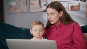 Happy young mother and little son are communicating with friends online, being lockdown. Mom and child are sitting on sofa, using laptop, speaking on video chat with relatives. 