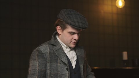 Mafia gangster in plaid suit in 1920s in room with yellow lamps undresses and takes off his classic plaid cap. The detective is in his office.