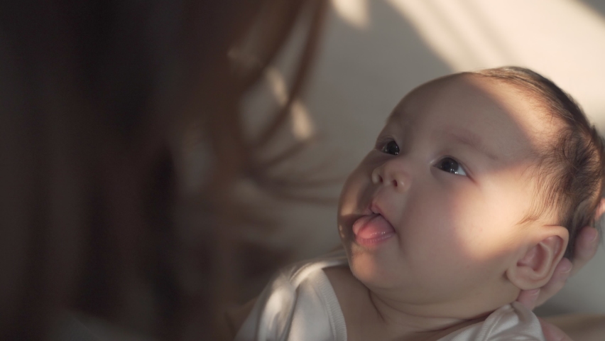 asian woman mother holding baby infant in arm playing and talking with kid on sofa couch in living room at home in evening. happy baby learning communication by smile, laugh, and Stick out tongue. Royalty-Free Stock Footage #1069129624