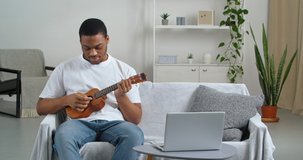 Afro american man wears white t-shirt and jeans sitting in cozy living room on couch watching music video lesson online using laptop learning to play small ukulele rejoices making victory hand gesture