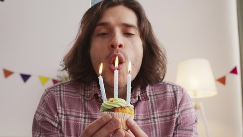 Camera pov of young man in party cap blowing candles on holiday cake, celebrating birthday online at home