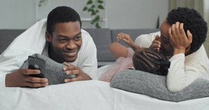 Afro american family three people lie on couch together, young father with mobile phone overhears secret conversation between mother and daughter, mom whispers information to little girl in ear