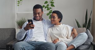 Loving afro american ethnic couple sitting on sofa at home talking spending time together at holidays. Mixed race smiled man and woman relaxing on couch with smartphone communication in social network