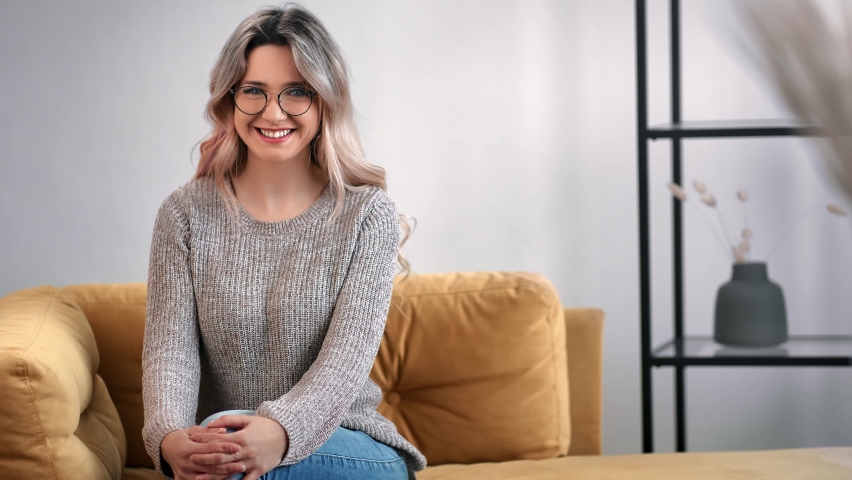 Cheerful young modern lady posing on comfortable yellow couch relaxing at living room having positive emotion. Happy beautiful blonde female in glasses sitting on sofa. Shot with RED camera in 4K