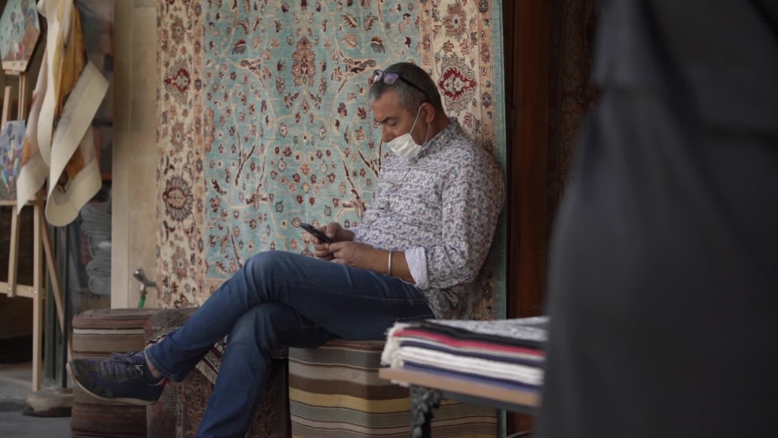 ISTANBUL, TURKEY - NOVEMBER 12 2020: Concentrated local man with black sunglasses and disposable face mask sits on chair near shop with carpets holding smartphone.