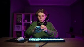 Young gamer in a headset sits at a computer at night in a room and taking selfie on smartphone camera, shows a gesture of peace.