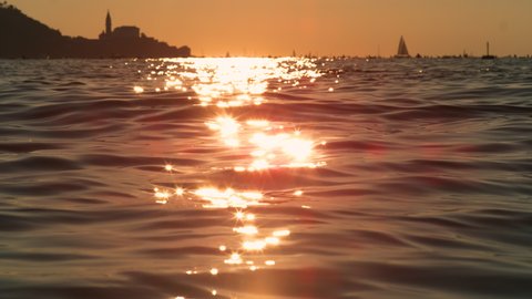 SLOW MOTION, LENS FLARE, CLOSE UP: Calm Adriatic sea near Piran glistens in the golden summer evening sunshine. Spectacular shot of the ocean and Piran cityscape in the distance at gorgeous sunrise.