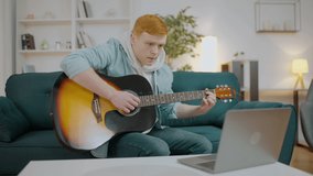 Young male learning to play acoustic guitar, watching online tutorial on laptop