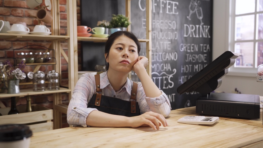Young female barman sitting leaning on wooden bar counter looking aside bored while waiting for customer in coffee shop. Beautiful asian barista in apron daydreaming with no client coming in cafe. Royalty-Free Stock Footage #1069159954