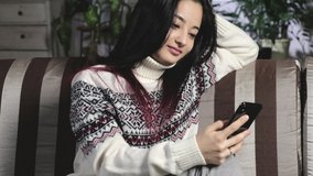 Attractive Asian girl with glasses, brunette in warm sweater and socks, sits relaxed on the couch, holds smartphone in her hands and surfs the Web in search of content. Online education in quarantine