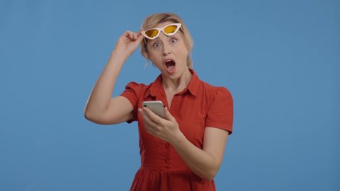 The woman is surprised at the news she sees while surfing the internet with her phone, and she lifts her glasses and looks at the camera. Portrait of surprised woman looking at camera.Slow motion vide