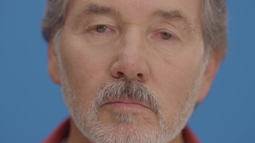 In front of the blue background, bearded old man looks ahead. Face plan. Close up.