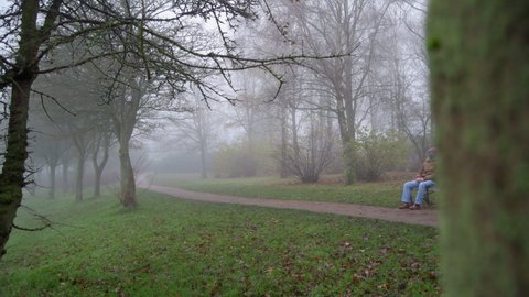 A wide shot of an elderly man as he sits by himself in a park on a foggy morning