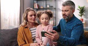 Cheerful positive Caucasian family with kid videochatting on mobile phone. Mother and father with cute small girl daughter speaking on video call online on cellphone and waving hand close up portrait