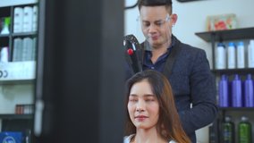 Asian professional male stylist using hair dryer blow woman's hair in salon. The man wearing mask and face shield to prevent from coronavirus infection during pandemic. Beauty salon business concept.