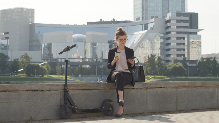 Full shot of young caucasian woman sitting on bridge by river in city centre on sunny day drinking coffee and using smartphone with electric scooter next to her