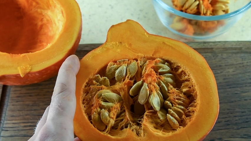 Cream of roasted pumpkin soup recipe. Step 2. Person removes seeds with spoon from orange pumpkin cut in half lying on cutting board. Point of view. Slow motion video. Home cooking theme.