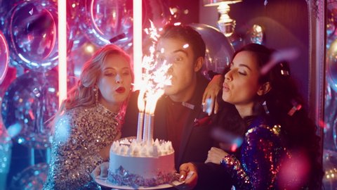 Joyful friends blowing candles together in nightclub. Young people holding birthday cake at party in night club. Sweet girls clapping hands at happy birthday party on neon lamps background.