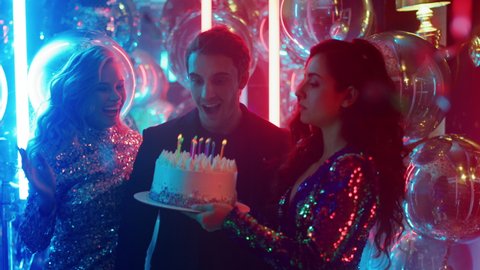 Happy friends congratulating guy in nightclub. Joyful man blowing candles at birthday party in night club. Attractive people celebrating happy birthday with cake on neon lamps background.