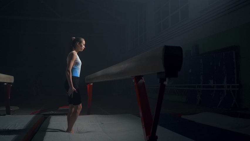 sporty teenager girl is training with balance beam in dark gymnastic hall, discipline of artistic gymnastics, slow motion shot