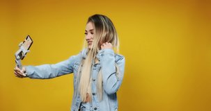 Young happy blonde girl teen blogger makes selfie on smartphone in wireless headphones, and smiles on yellow background summer. Emotions of people. Relax. 