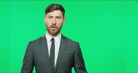 Caucasian presenter in business suit with tie tells news block sitting in studio on green screen background.