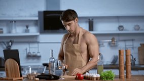 shirtless man in apron cooking in kitchen, looking at camera and smiling