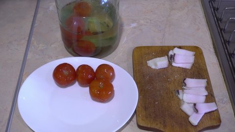 Hand cuts a large piece of salted lard into small pieces with a knife on a wooden board, canned tomatoes in a jar