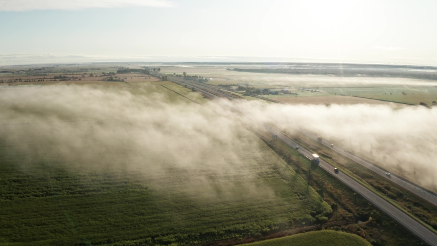 Aerial view of fog hanging low over green farmland cut by highway with traffic moving both directions. Morning landscape scenery in the plains with low clouds over agricultural fields. Drone descends.