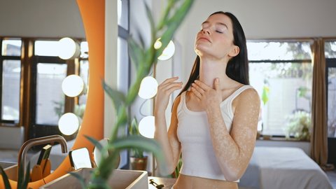 Young woman with vitiligo applying pampering serum on her neckline and shoulders skin, standing near mirror at bathroom